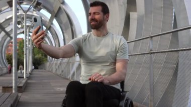 Tekerlekli sandalyedeki mutlu orta yaşlı adam bir video görüşmesi yapıyor, modern erişilebilir kentsel alanlar sergiliyor..