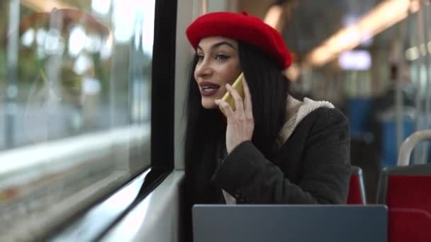 赤いベレーのファッショナブルな若い女性は 電車の窓に座っている間 彼女のスマートフォンで話します 会話に従事 列車で電話をするスタイリッシュな若い女性 — ストック動画