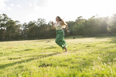 Saf bir neşe anı yakalayan genç bir kadın, güneşli bir tarlada çıplak ayakla koşuyor, yeşil eteği ve saçları rüzgarda uçuşuyor..