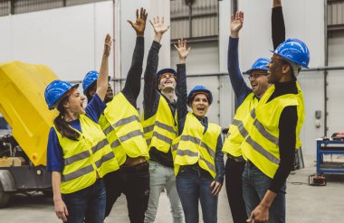 Güvenlik teçhizatlı bir grup mühendis, endüstriyel bir fabrikada ellerini kaldırarak kutlama yapıyorlar..