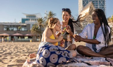 Bikinili ve beyaz gömlekli üç genç kadın sahilde otururken gülüyor ve meyve bardaklarını paylaşıyorlar..