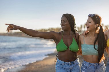Bikinili ve kot şortlu iki genç kadın kumsalda yürüyor, gülümsüyor ve uzaktaki bir şeyi işaret ediyor..