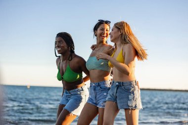 Bikinili ve kot şortlu üç genç kadın gülüyor ve kumsalda yürüyor güneşli bir günün tadını çıkarıyorlar..