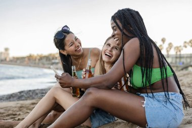 Bikinili ve kot şortlu üç genç kadın sahilde birlikte gülüp fotoğraf çekiyorlar, içkiden keyif alıyorlar..