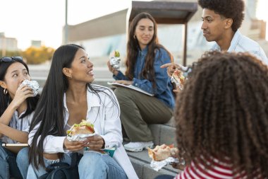 Güneşli bir kampüs gününde elinde defteriyle bir arkadaşla gülüp sohbet eden bir üniversite öğrencisi..