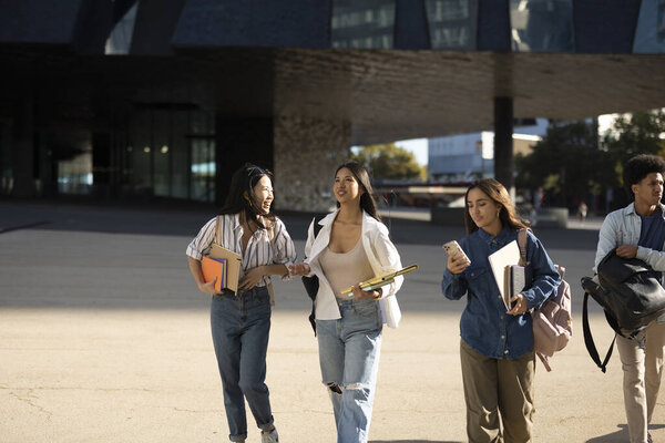 Группа студентов различных колледжей, гуляющих и болтающих по кампусу, с книгами и рюкзаками.