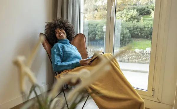 stock image Young woman in blue sweater and yellow blanket relaxing and reading a book by a large window.