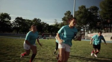 Kırmızı ve mavi formalı bayan rugby oyuncuları yoğun bir mücadele içinde, bir maç sırasında rugby topuna odaklanıyorlar..
