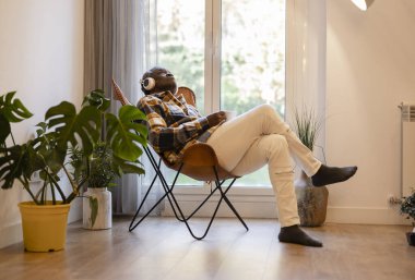 Yaşlı bir adam bir sandalyede oturuyor, kulaklıklarla dinleniyor ve rahat bir evde kahve içiyor..