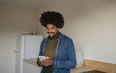Modern bir mutfakta akıllı telefonunu kullanırken gülümseyen kıvırcık saçlı genç Afrikalı Amerikalı adam..