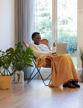 Genç Afrikalı Amerikalı bir kadın kahvesini içiyor ve laptopuyla çalışıyor, pencerenin kenarında oturmuş, etrafında bitkilerle..