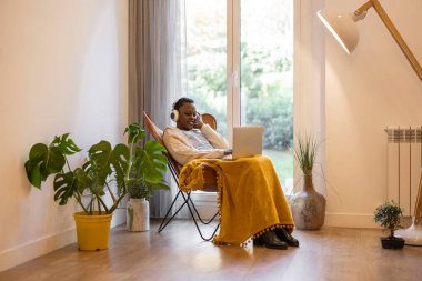 Genç Afrikalı Amerikalı kadın bilgisayarını kullanıyor ve kulaklık takıyor. Pencere kenarında bitkilerle rahatça oturuyor..