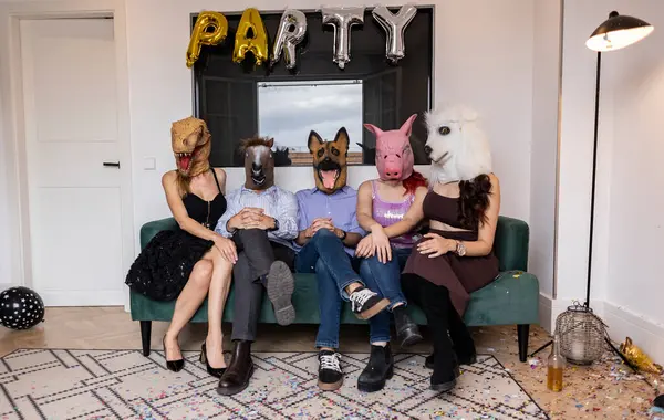 stock image Group of friends in their 30s wearing various animal masks, sitting on a sofa at a themed party with confetti and balloons.