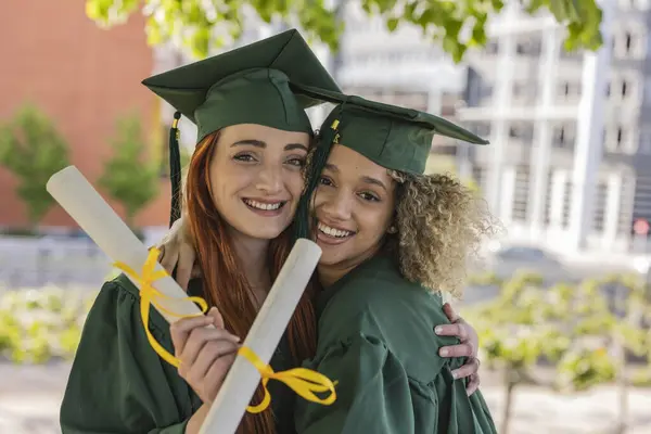 Stock image Red-haired woman and mixed-race woman in green graduation gowns, smiling and embracing while holding their diplomas.