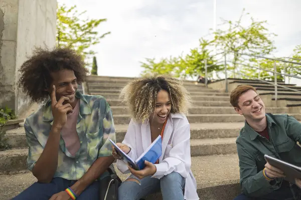 Basamaklarda oturan üç farklı genç yetişkin, kitaplar ve bir tabletle birlikte gülüyor ve ders çalışıyorlar..