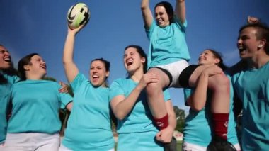 Heyecanlı bayan rugby oyuncuları, elinde kupa olan bir takım arkadaşını kaldırıp zaferlerini neşeli ifadelerle kutluyorlar..