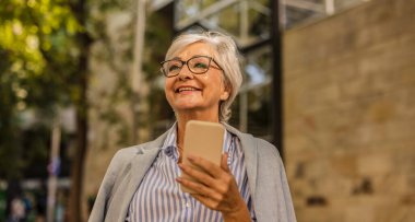 Güneşli bir günde dışarıda akıllı telefonunu kullanırken gülümseyen yaşlı bir kadın..