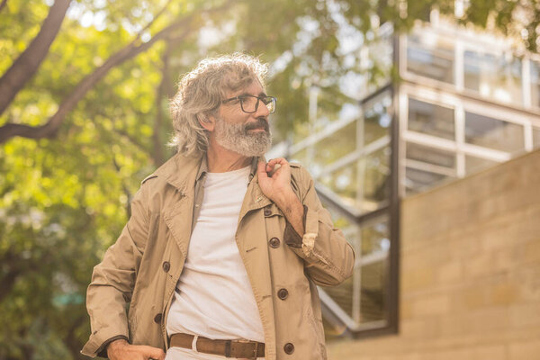 Elderly man with glasses and a trench coat enjoying a sunny day in the city.