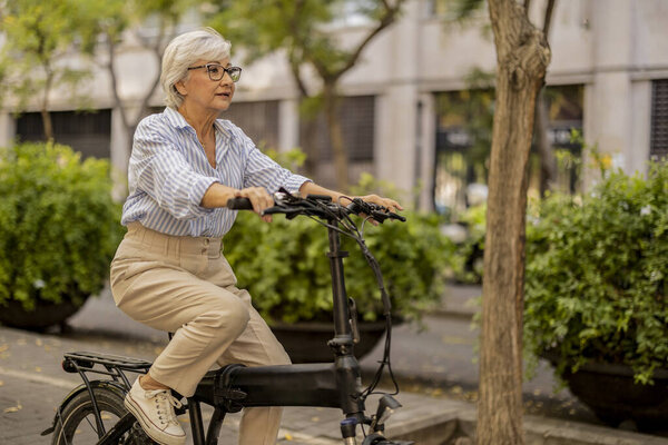 Elderly woman with glasses riding a bike through an urban park on a sunny day.