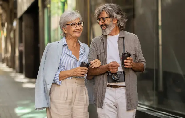 stock image Elderly couple smiling, holding coffee cups, and walking arm-in-arm on a sunny day.