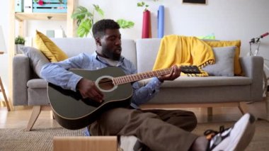 Günlük kıyafetli bir adam yerde oturuyor ve rahat bir oturma odasında akustik gitar çalıyor..