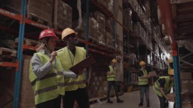 Çok ırklı depo çalışanları, güvenlik ekipmanları giyip envanteri tartışırken panoya bakıyorlar. Dolu depo ortamı.