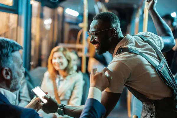 stock image Smiling worker showing something on his phone to a fellow traveler on a night bus.