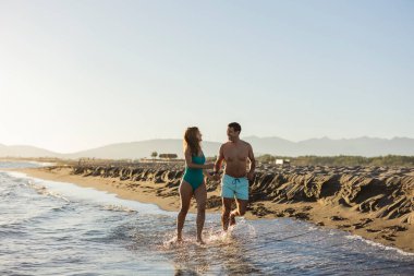 Gülümseyen çift el ele tutuşuyor ve güneşli bir sahilde kıyı boyunca koşuyor..