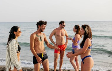Bir grup genç yetişkin sahilde gülüyor ve sohbet ediyor, güneşli bir günün tadını çıkarıyorlar..