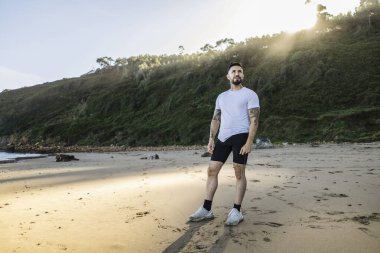 Spor giyim sektöründe genç bir adam arka planda yemyeşil tepelerle güneşli bir plajda dikiliyor..