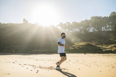 Güneşli bir sabah kumlu bir plajda koşan atletik bir adam, doğa ile çevrili..
