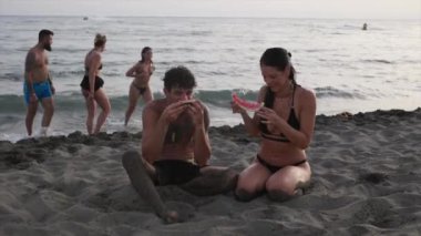 Bir grup genç yetişkin sahilde oturmuş, birlikte karpuz dilimleri yiyorlar..