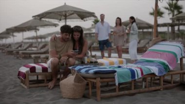 Bir grup arkadaş plaj şezlonglarında dinleniyor, bir çift telefonu kontrol ediyor ve diğerleri arka planda sohbet ediyor..