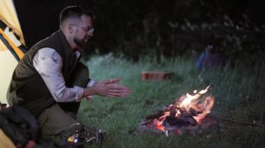 30 'lu yaşlarda bir adam gece kamp ateşinde ellerini ısıtıyor ve huzurlu bir kamp deneyimi yaşıyor..
