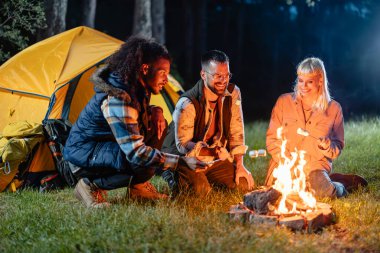 Bir grup arkadaş, geceleyin ormanda çadırın yakınındaki bir kamp ateşinde lokum kızartıyorlar..
