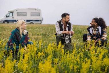 Arka planda bir karavanla sarı çiçeklerle dolu bir alanda oturup sohbet eden üç arkadaş..