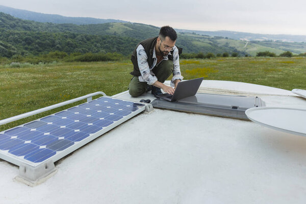 Man using a laptop on the roof of an RV equipped with solar panels, surrounded by scenic nature.
