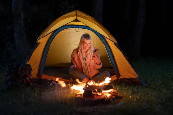 stock image Woman sitting in a tent by a campfire at night, smiling while looking at her smart phone.