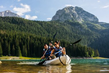 Genç bir çift, yoğun orman ve yüksek dağlarla çevrili, berrak bir dağ gölünde kayak yapıyor, can yelekleri giyiyor ve maceranın tadını çıkarıyor..