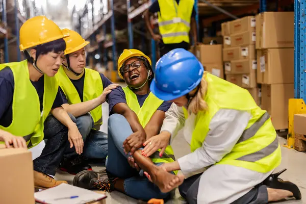 Kadın depo çalışanı, yaralanma sonrası meslektaşlarından ilk yardım alıyor, işyeri güvenliğini vurguluyor.
