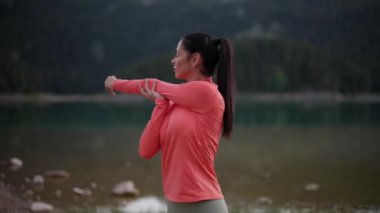 Mercan kaplı bir kadın kollarını dingin bir dağ gölünün yakınına uzatıyor..