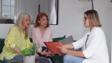 Bir ofis ortamında kadın bir profesyonel tarafından sunulan belgeyi dikkatlice inceleyen ve imzalayan iki olgun kadın..