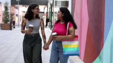 Latin Amerikalı lezbiyen çift kahve fincanlarıyla yürürken birbirlerine gülümsüyor, biri gökkuşağı çizgili bir çanta tutuyor..
