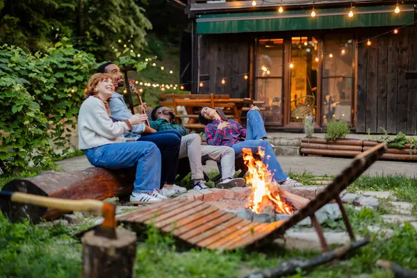 stock image A group of multiracial friends relax around a campfire, laugh and enjoy the cozy atmosphere of a rustic outdoor setting, illuminated by string lights.