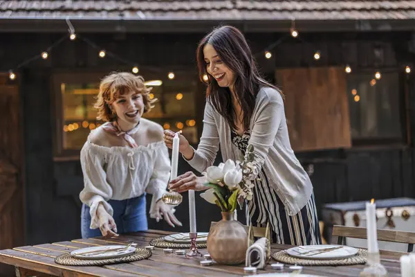 stock image Two women in their 30s happily setting up a candlelit outdoor dinner table, preparing for an intimate evening gathering.