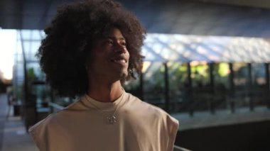 Açık hava ortamında kendine güvenen kıvırcık saçlı, bej gömlekli ve kolyeli genç Afro-Amerikan bir adam..