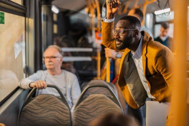 Orta yaşlı iş adamı dizüstü bilgisayar tutuyor ve bir otobüs dolusu çok jenerasyonlu insanın yanında oturan diğer yolcularla birlikte ayrılmaya hazırlanıyor..