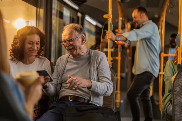 stock image Elderly man and woman smiling and enjoying a conversation while looking at a smart phone on a public bus during a sunny day.