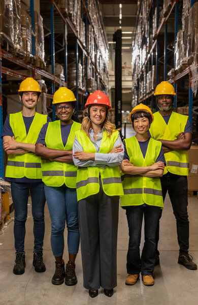 A group of five diverse workers wearing hard hats and safety vests in a warehouse, standing confidently.