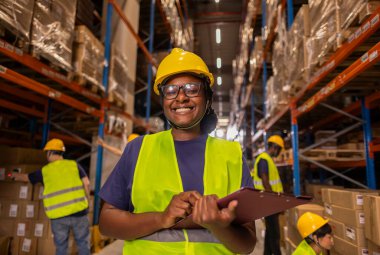 A smiling African warehouse worker in safety gear holding a clipboard while standing in a busy warehouse aisle. clipart